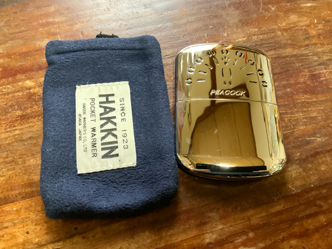 ハクキンカイロの本体(真鍮性で掌サイズ)と布製のケース(商品名が入ったラベルが縫い付けてあります)